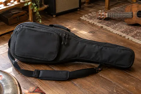 Housse noire Kala pour ukulélé concert, avec poche extérieure et bandoulière réglable, posée sur un sol de bois
