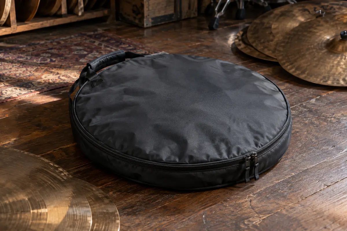 Housse ronde en nylon gris sombre pour cymbales, posée sur un sol de bois patiné près de cymbales
